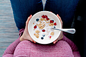 Top view of crop unrecognizable female with bowl of cornflakes and ripe berries with milk for breakfast in light room