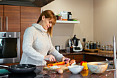Smiling adult female in sweater peeling carrots wooden cutting board at counter with built in stovetop and bowls while making dinner