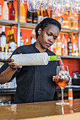 Focused African American female barkeeper pouring alcohol in glass with ice cube while making cocktail in bar