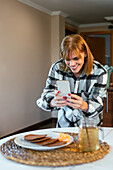 Smiling adult lady in checkered shirt standing near table and taking photo of pancakes and glass mug of drink with phone