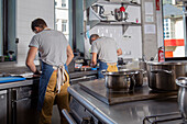 Back view of young Hispanic male cook in casual clothes and apron cutting sweet potato while working in restaurant kitchen near colleague