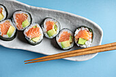 Leckere norwegische Futomaki-Sushi-Rollen mit Lachs und Avocado, serviert mit Stäbchen auf blauem Hintergrund in einem hellen Studio