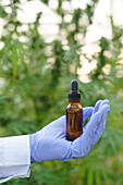 Hand of crop scientist wearing white robe and latex gloves demonstrating glass bottle with liquid drug