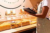 Cropped side view of unrecognizable black female vendor in uniform with tablet smiling and inspecting pasta in glass container during work in zero waste grocery store looking away