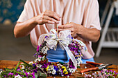 Cropped unrecognizable female florist making flower wreath with ribbon bow while working in floral shop