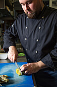 Bearded male chef cutting fresh artichokes while preparing delectable dish on table in kitchen of contemporary restaurant