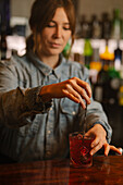 Focused female barkeeper mixing with spoon alcoholic red old fashioned cocktail with ice cubes while standing at bar counter during work