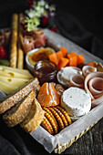 From above brunch box with assorted sliced meats various types of cheese and crispbreads arranged near ripe cup kiwi sweet strawberries and peeled mandarin near jam in glass jar on wooden table near napkin