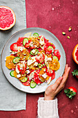 Overhead of crop person eating delicious healthy salad with strawberry and orange and cucumber slices with cheese and pistachios on plate
