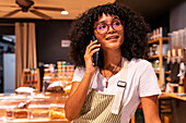 Glad young African American woman in apron and glasses leaning on counter and talking on cellphone while working in zero waste grocery store