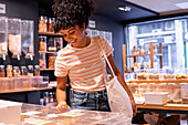 Cheerful young African American woman in casual clothes with curly hair smiling and using tote bag while visiting eco friendly grocery shop