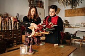 Fröhliche Freundinnen stehen am Tisch mit Kerzen und basteln kreative Weihnachtssträuße für die Weihnachtsfeier