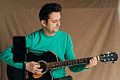 Adult male vlogger with guitar and plectrum recording video tutorial on cellphone at home
