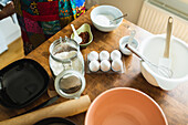 From above set of various kitchen utensils and fresh eggs sugar and flour prepared for baking and placed on wooden kitchen table with anonymous person wearing apron in bright daylight