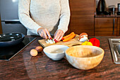 Faceless lady in sweater dicing onion on cutting board with peeled carrot while standing at counter with stovetop and bowls