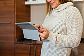 Crop smiling adult lady in cozy sweater tapping screen of tablet while standing against cabinets in kitchen at home