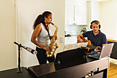 Hispanic Frau mit langen lockigen Haaren stehen und spielen Saxophon in der Nähe von positiven männlichen in Kopfhörer sitzen und spielen akustische Gitarre in hellen Raum in der Tageszeit