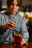 Blurred unrecognizable barkeeper adding lemon zest in glass with alcoholic red old fashioned cocktail while working in bar on blurred background