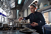 Woman in dark uniform putting shrimp into saucepan with hot oil while working in kitchen of modern restaurant