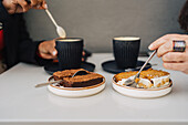 Crop anonymous female eating sweet cake with black friend stirring hot cappuccino with spoon in coffee shop