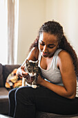 Calm Hispanic woman with long curly hair sitting on soft sofa and stroking domestic cat at home in daytime