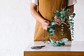 Crop unrecognizable female florist in apron creating composition with fresh green Eucalyptus leaves standing at table in light studio