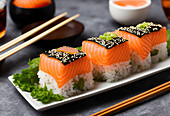 Appetizing sushi rolls with rice and salmon served on plate with green salad leaves and sesame seeds placed near chopsticks against bowls of sauce