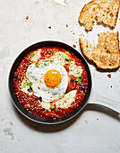 Draufsicht auf eine Bratpfanne mit appetitlicher Shakshuka mit Ei und Käse neben Brot auf dem Tisch