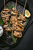 From above of appetizing freshly cooked meat on skewers served on tray on black table with bowl of sauce