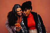 Hispanic lady and African American female friend looking at screen of mobile phone happily while standing against red backdrop