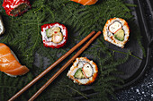 Tasty fresh assorted sushi served on green plant twigs on black plate with soy sauce on marble table near chopsticks