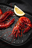 Delicious cooked red prawns on tray with coarse salt and juicy lemon pieces on dark background