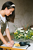 Side view of concentrated adult ethnic female floral designer with long dark hair in apron serving table with plates and cutlery and decorating with fresh delicate flowers