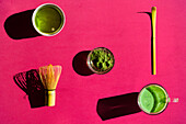 Blick von oben auf einen hellen Hintergrund mit einem Glas grünem Tee und getrocknetem Matcha mit Bambus-Chasen und Chashaku auf einer rosa Fläche