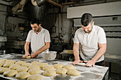 Älterer bärtiger ethnischer Bäcker formt Brot aus Teig am Tisch mit Mehl und Schüssel in einer Bäckerei