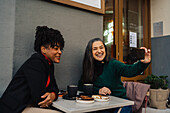 Positive ethnic mature woman having latte and taking self portrait with laughing African American girlfriend in outdoor coffee house