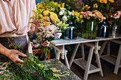 Crop faceless female florist creating flower bouquet while working at table in floral store