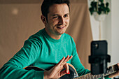 Crop cheerful adult male vlogger with guitar and plectrum recording video tutorial on cellphone at home