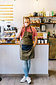 Full body of serious male barista in apron looking at camera while drinking hot coffee near counter in modern coffee house