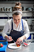 Smiling woman in apron using spoon to arrange crumbs and strawberry puree on plate during dessert preparation in restaurant