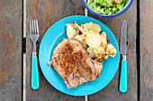 Draufsicht auf ein appetitliches gebratenes Steak mit Kartoffeln, serviert auf einem blauen Teller mit Gabel und Messer neben einer Schüssel mit Salat auf einem Holztisch