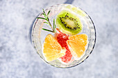 Top view of summer tropical cocktail of vodka and sprite with ripe fruits and rosemary on grey background