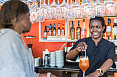 Cheerful black female barkeeper serving glass of refreshing cocktail for client in bar