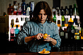 Focused female bartender taking off lemon zest while preparing for making cocktail in bar with various alcohol drinks during work