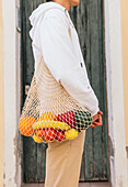 Side view of cropped unrecognizable male standing with eco friendly mesh bag full of fresh fruits in city street
