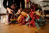 Crop unrecognizable female florist standing and cutting twigs of fir while arranging Christmas bouquet on wooden table