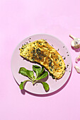 Tasty omelette on plate against fresh parsley sprigs with garlic cloves on pink background