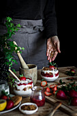 Erntehelferin in schwarzem Hemd und grauer Schürze beim Kochen eines leckeren Desserts mit Käse und frischen reifen Erdbeeren und Keksen am Tisch vor schwarzem Hintergrund