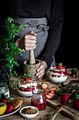 Crop Person in schwarzem Hemd und grauer Schürze mit Mörser beim Kochen köstlichen Dessert mit Käse und frischen reifen Erdbeeren und Kekse am Tisch vor schwarzem Hintergrund
