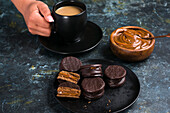 Chocolate alfajores with dulce de leche placed on black plate against blurred background of crop person holding cup